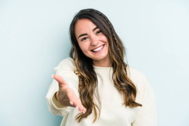 Mavi arka planda izole edilmiş genç beyaz kadın selamlama jesti olarak kameraya elini uzatıyor..