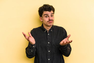 Sarı arka planda izole edilmiş genç beyaz adam sorgulama sırasında omuz silkiyor ve kuşkulanıyor..