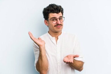 Kafkasyalı genç adam mavi arka planda izole edilmiş. Kafası karışmış ve omuz silkiyor. Kopya alanını tutmak için..