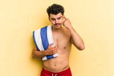 Young caucasian man holding beach towel isolated on yellow background pointing temple with finger, thinking, focused on a task.