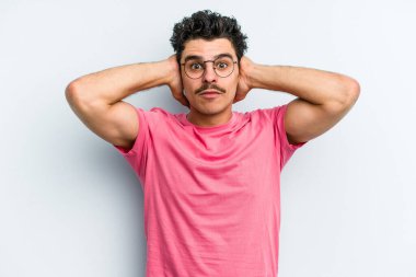 Kafkasyalı genç adam mavi arka planda izole edilmiş kulakları kulaklarıyla kapatıyor çok fazla ses duymamaya çalışıyor..