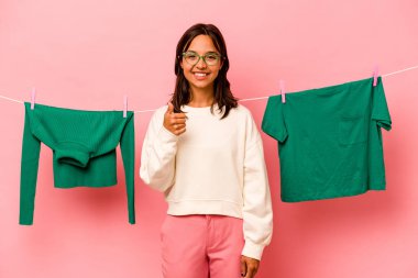 Çamaşır sepetini tutan genç İspanyol kadın pembe arka planda izole edilmiş gülümsüyor ve başparmağını kaldırıyor.