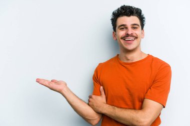 Mavi arka planda izole edilmiş genç beyaz bir adam bir palmiye üzerinde fotokopi alanı ve belinde bir el daha gösteriyor..