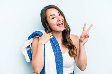 Young caucasian woman holding beach towel isolated on blue background joyful and carefree showing a peace symbol with fingers.