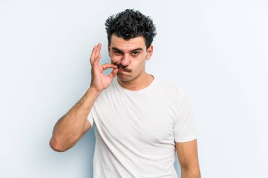 Genç beyaz adam mavi arka planda izole edilmiş ve parmakları dudaklarında bir sır saklıyor..