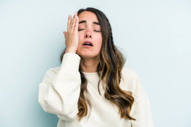 Mavi arka planda izole edilmiş genç beyaz kadın yorgun ve uykulu ellerini başının üstünde tutuyor..