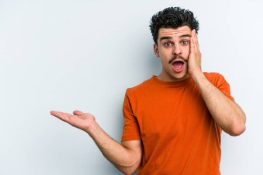 Mavi arka planda izole edilmiş genç beyaz adam, avuç içi fotokopi alanını elinde tutmaktan etkilenmiş..