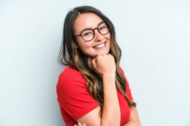 Mavi arka planda izole edilmiş genç beyaz kadın mutlu ve kendinden emin gülümsüyor, elleriyle çenesine dokunuyor..