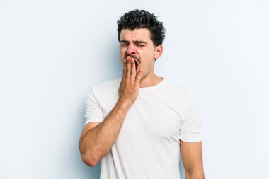 Mavi arka planda izole edilmiş genç beyaz adam esniyor. Ağzı eliyle kapatan yorgun bir hareket sergiliyor..