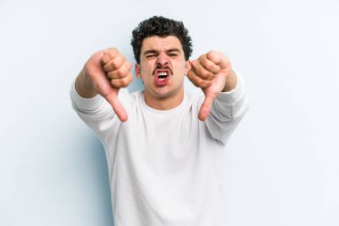 Mavi arka planda izole edilmiş genç beyaz bir adam baş parmağını aşağı gösteriyor ve hoşlanmadığını ifade ediyor..