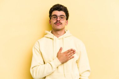 Sarı arka planda izole edilmiş genç beyaz adam yemin ediyor, elini göğsüne koyuyor..