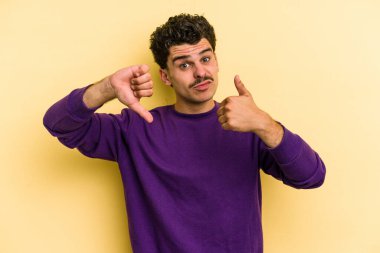 Sarı arka planda izole edilmiş genç beyaz adam baş parmağını kaldırıyor ve baş parmak aşağı, zor seçim konsepti.