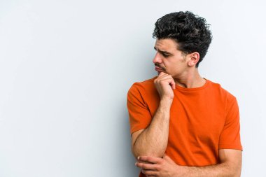 Mavi arka planda izole edilmiş genç beyaz adam şüpheli ve şüpheci bir ifadeyle yana bakıyor..