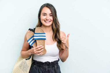 Young caucasian woman holding sandals isolated on blue background smiling and raising thumb up