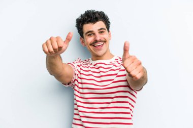 Kafkasyalı genç adam mavi arka planda izole edilmiş. Baş parmağını kaldırıyor, gülümsüyor ve kendinden emin..