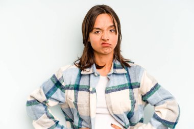 Genç İspanyol kadın mavi arka planda izole edilmiş. Mutsuz ve alaycı bir ifadeyle kameraya bakıyor..