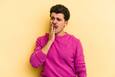 Sarı arka planda izole edilmiş genç beyaz adam esniyor. Ağzı eliyle kapatan yorgun bir hareket sergiliyor..