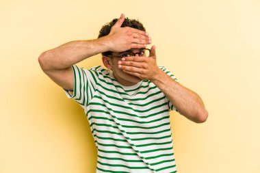 Sarı arka planda izole edilmiş genç beyaz adam kameraya parmaklarıyla göz kırpıyor, yüzünü kapatmaktan utanıyor..