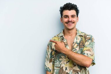 Mavi arka planda izole edilmiş genç beyaz adam gülümsüyor ve boş alanda bir şeyler gösteriyor..