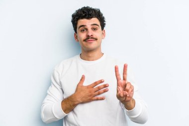 Kafkasyalı genç adam mavi arka planda izole edilmiş yemin ediyor, elini göğsüne koyuyor..