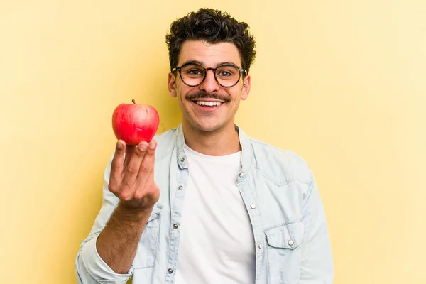Young caucasian man holding an apple isolated on yellow background happy, smiling and cheerful.