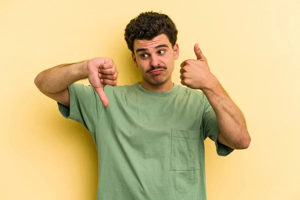 Young caucasian man isolated on yellow background showing thumbs up and thumbs down, difficult choose concept