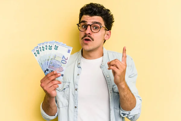 Young caucasian man holding banknotes isolated on yellow background having some great idea, concept of creativity.