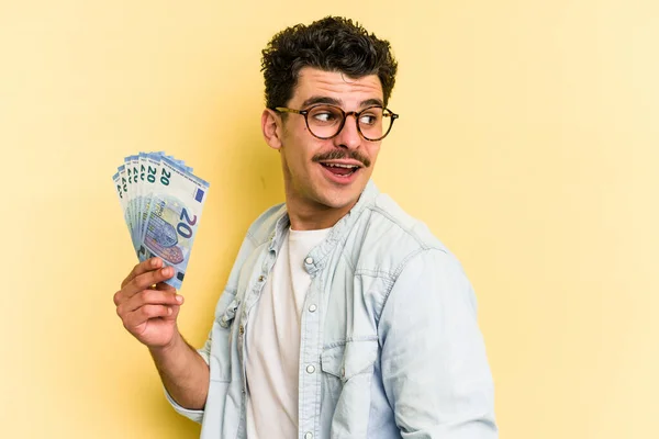 Young caucasian man holding banknotes isolated on yellow background looks aside smiling, cheerful and pleasant.