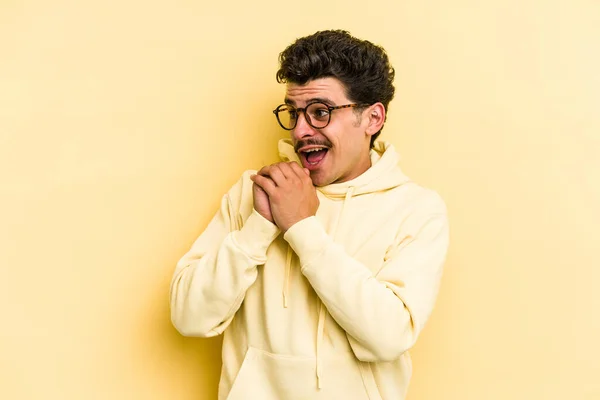 Young caucasian man isolated on yellow background praying for luck, amazed and opening mouth looking to front.