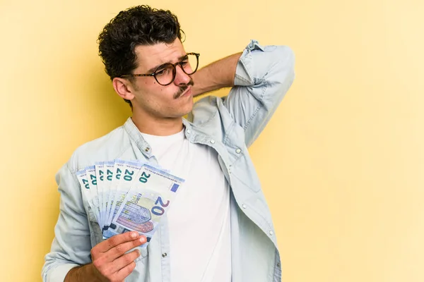 Young caucasian man holding banknotes isolated on yellow background touching back of head, thinking and making a choice.