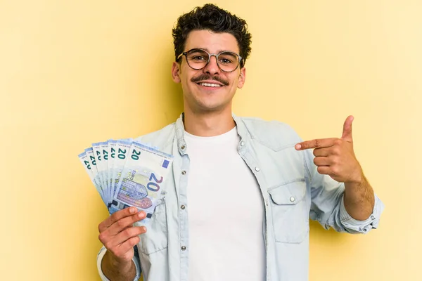 Young caucasian man holding banknotes isolated on yellow background person pointing by hand to a shirt copy space, proud and confident