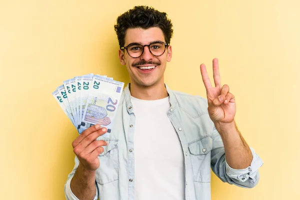 Young caucasian man holding banknotes isolated on yellow background joyful and carefree showing a peace symbol with fingers.