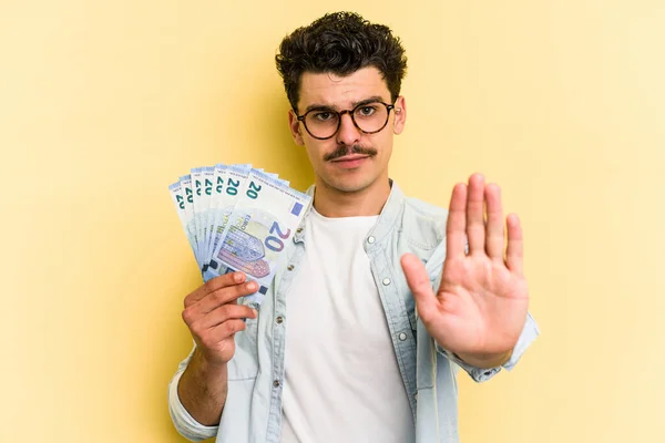 Young caucasian man holding banknotes isolated on yellow background standing with outstretched hand showing stop sign, preventing you.
