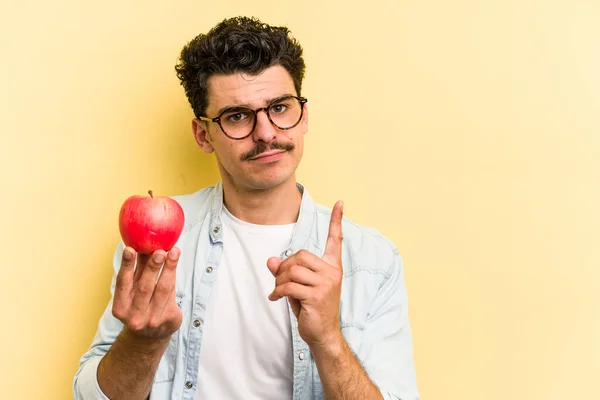 Young caucasian man holding an apple isolated on yellow background showing number one with finger.