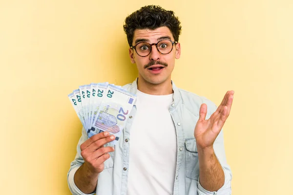 Young caucasian man holding banknotes isolated on yellow background surprised and shocked.