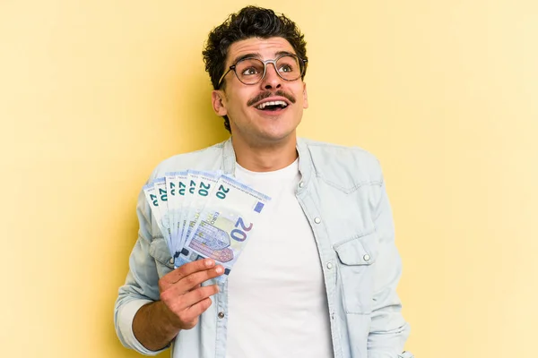 Young caucasian man holding banknotes isolated on yellow background dreaming of achieving goals and purposes