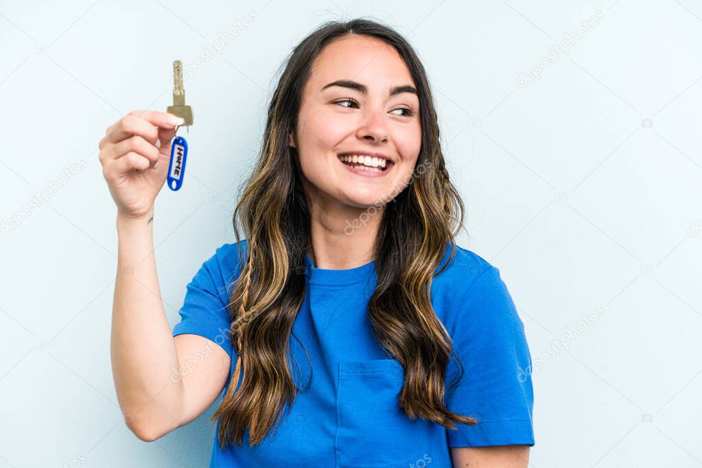 Young caucasian woman holding home keys isolated on blue background looks aside smiling, cheerful and pleasant.
