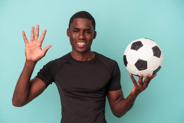 Mavi arka planda izole bir şekilde futbol oynayan genç Afrikalı Amerikalı beş numarayı parmaklarıyla gülümsüyor..