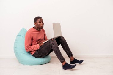Genç Afrikalı Amerikalı bir adam, beyaz arka planda izole edilmiş dizüstü bilgisayar kullanarak bir puf üzerinde oturuyor omuz silkiyor ve gözleri açık..
