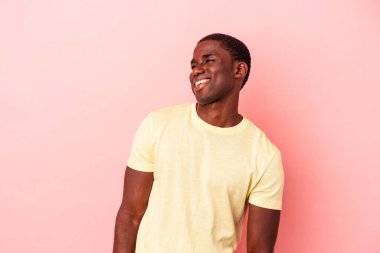 Genç, Afro-Amerikalı bir adam pembe arka planda izole edilmiş rahat ve mutlu bir şekilde gülüyor, boynunda dişlerini gösteriyor..