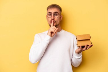 Sarı arka planda izole edilmiş bir burger tutan genç beyaz bir adam ya sır saklıyor ya da sessizlik istiyor..