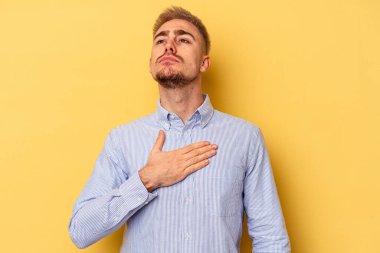 Sarı arka planda izole edilmiş genç beyaz adam yemin ediyor, elini göğsüne koyuyor..