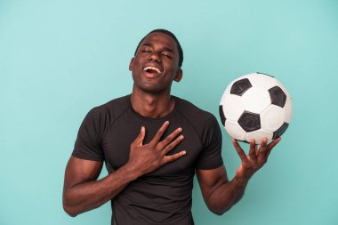 Mavi arka planda izole bir şekilde futbol oynayan genç Afrikalı Amerikalı adam elini göğsüne koyduğunda kahkahalar atıyor..