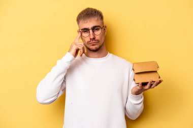 Elinde bir burger tutan genç beyaz adam sarı arka planda izole edilmiş, parmağıyla şakağı işaret eden, düşünen, bir göreve odaklanmış..