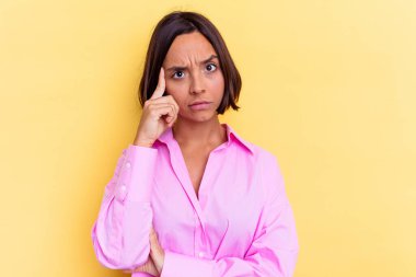 Sarı arka planda izole edilmiş genç melez bir kadın şakağı parmağıyla işaret ediyor, düşünüyor ve bir göreve odaklanıyor..