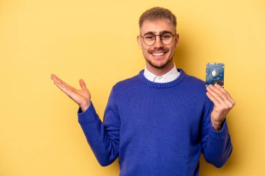 Young computer man isolated on yellow background showing a copy space on a palm and holding another hand on waist.