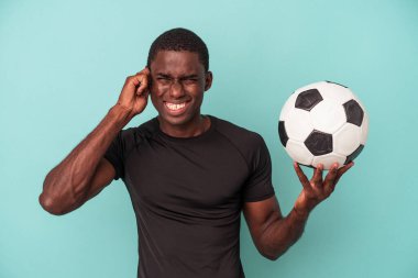 Futbol oynayan genç Afrikalı Amerikalı adam mavi arka planda izole edilmiş kulakları elleriyle kapatıyor..