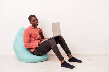 Genç Afro-Amerikalı bir adam beyaz arka planda izole edilmiş bir dizüstü bilgisayar kullanarak bir puf üzerinde oturuyor. Sanki sizi davet ediyormuş gibi..