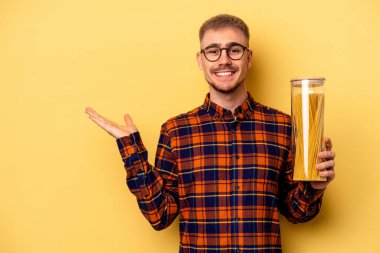 Young caucasian man holding spaghettis jar isolated on yellow background showing a copy space on a palm and holding another hand on waist.