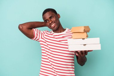 Elinde pizza ve burger tutan genç Afrikalı Amerikalı adam mavi arka planda izole edilmiş kafasının arkasına dokunuyor, düşünüyor ve bir seçim yapıyor..